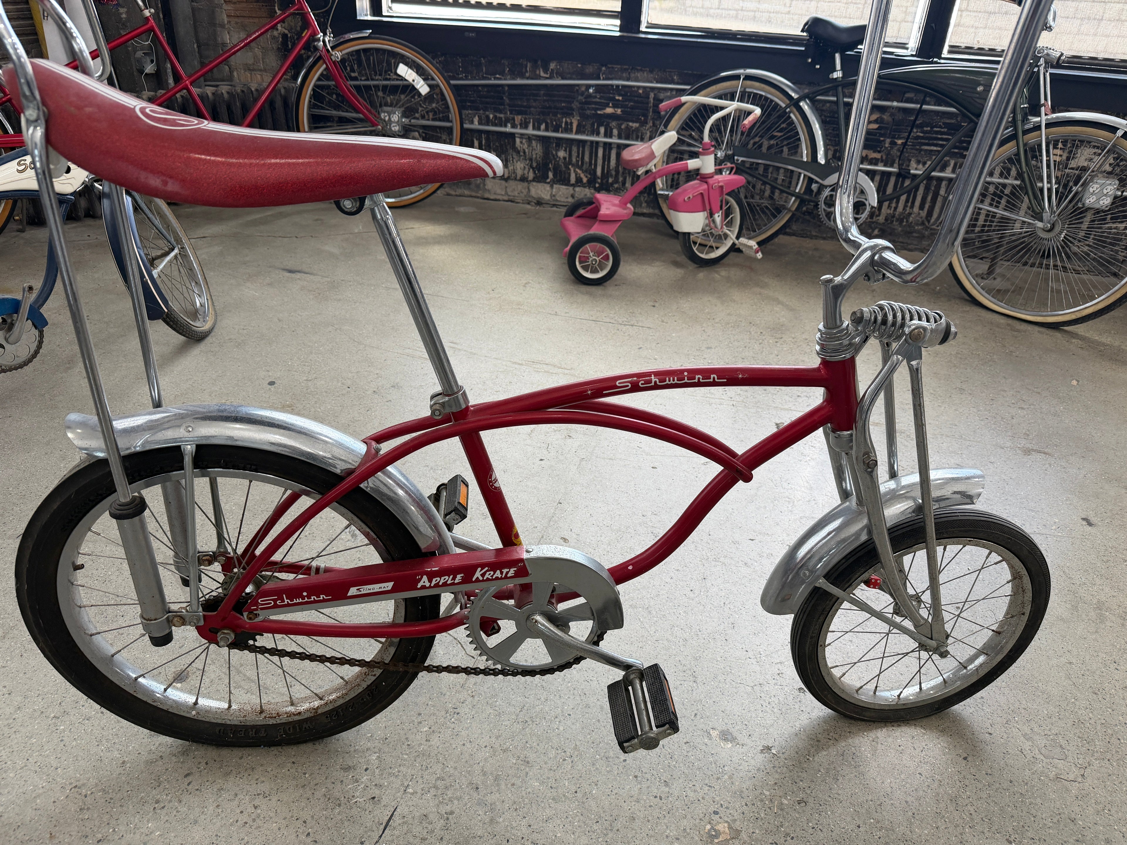 Vintage Schwinn Sting-Ray Apple Krate bicycle in red with banana seat, tall sissy bar, chrome fenders, springer-style front fork, and high-rise handlebars — full side profile at The Foundry by History Loves Company in downtown Pontiac, Michigan