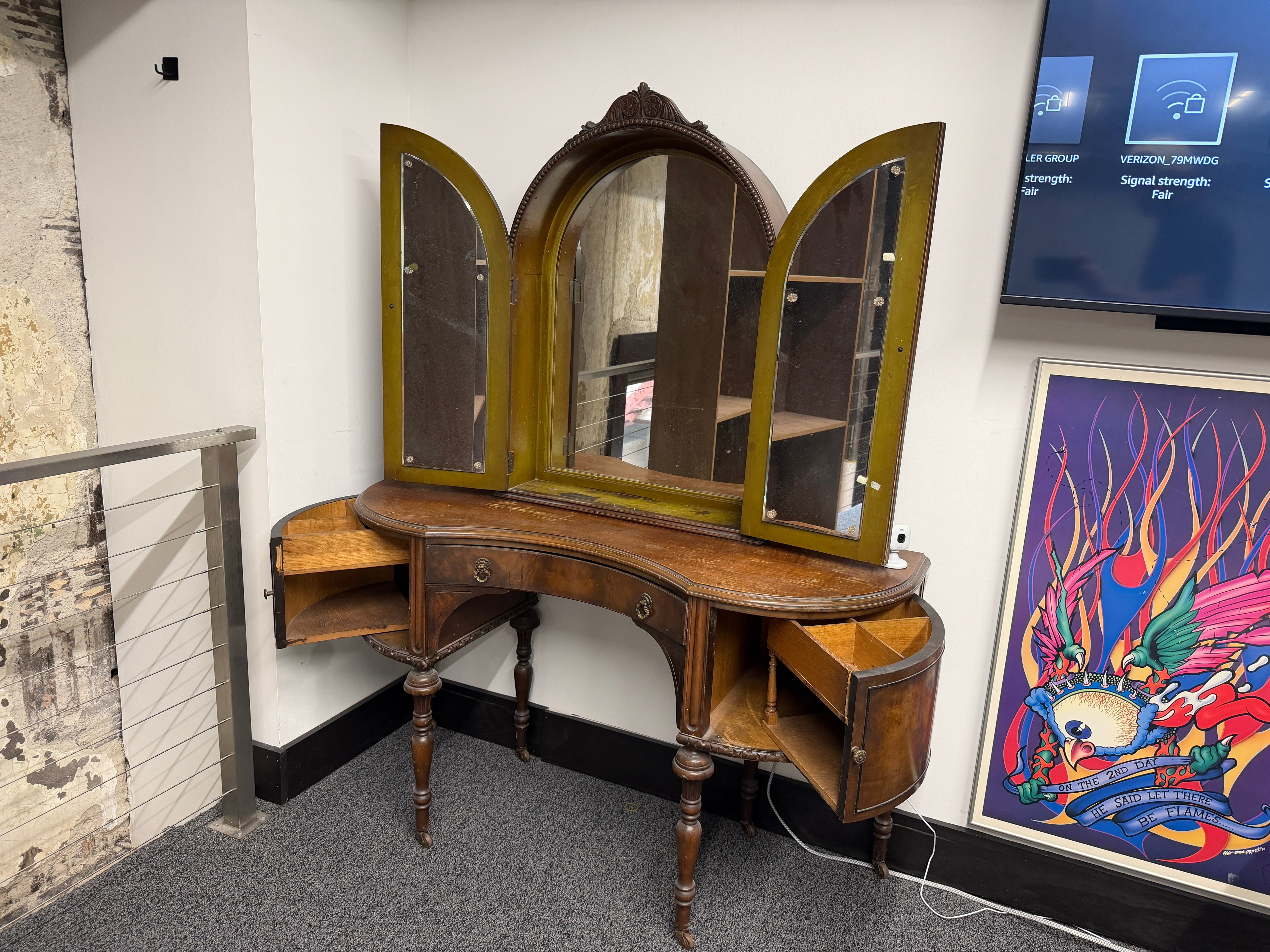 Antique tri-fold vanity dressing table with carved arched center mirror, folding side mirror doors, curved top surface, turned wood legs, side storage cabinets, and warm aged wood finish — on consignment at The Foundry by History Loves Company in downtown Pontiac, Michigan