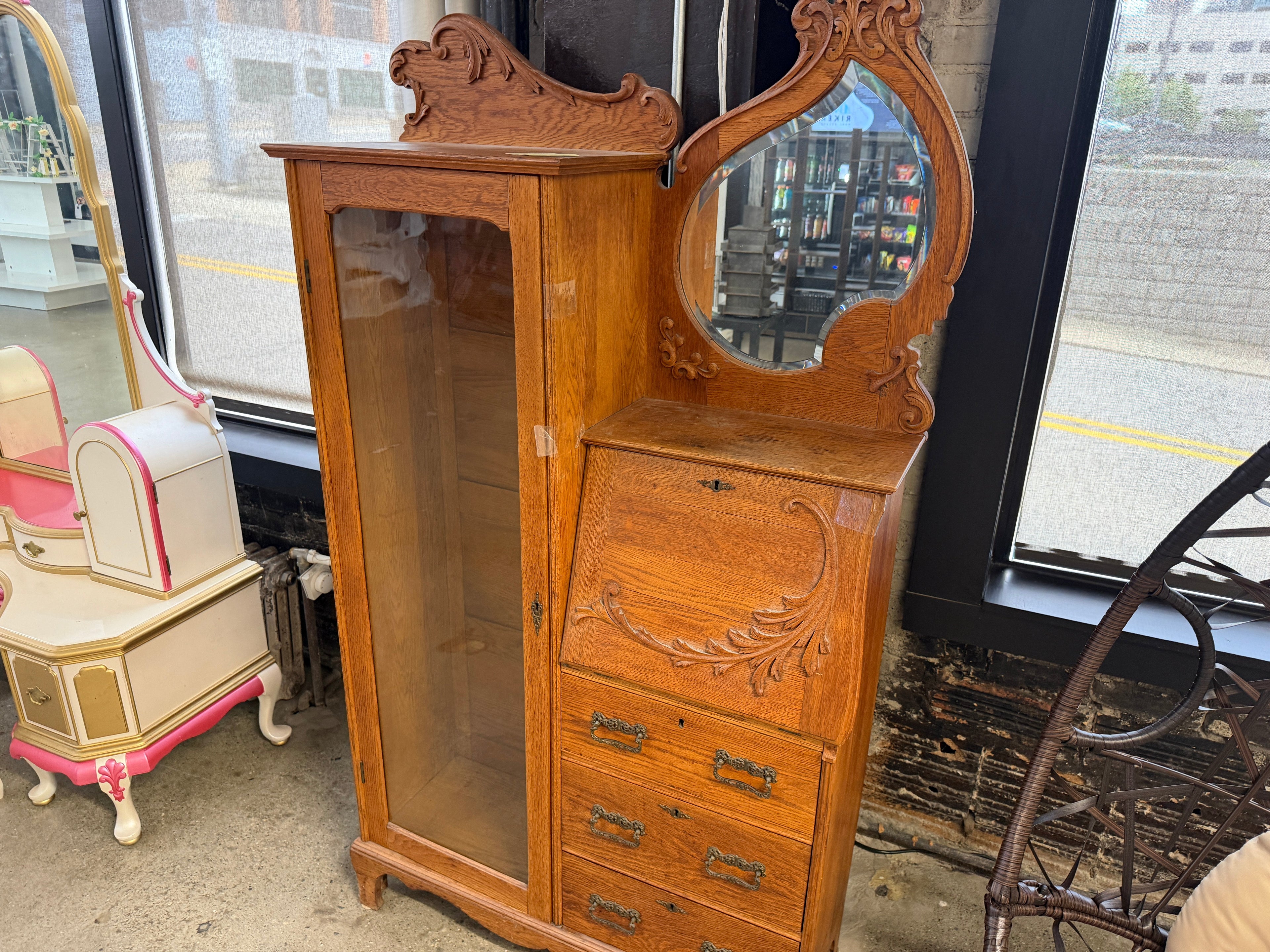 Antique oak side-by-side secretary bookcase with carved decorative crest, glass-front display cabinet, drop-front writing desk, oval mirror, lower drawers with brass hardware, and warm quarter-sawn oak grain — on consignment at The Foundry by History Loves Company in downtown Pontiac, Michigan