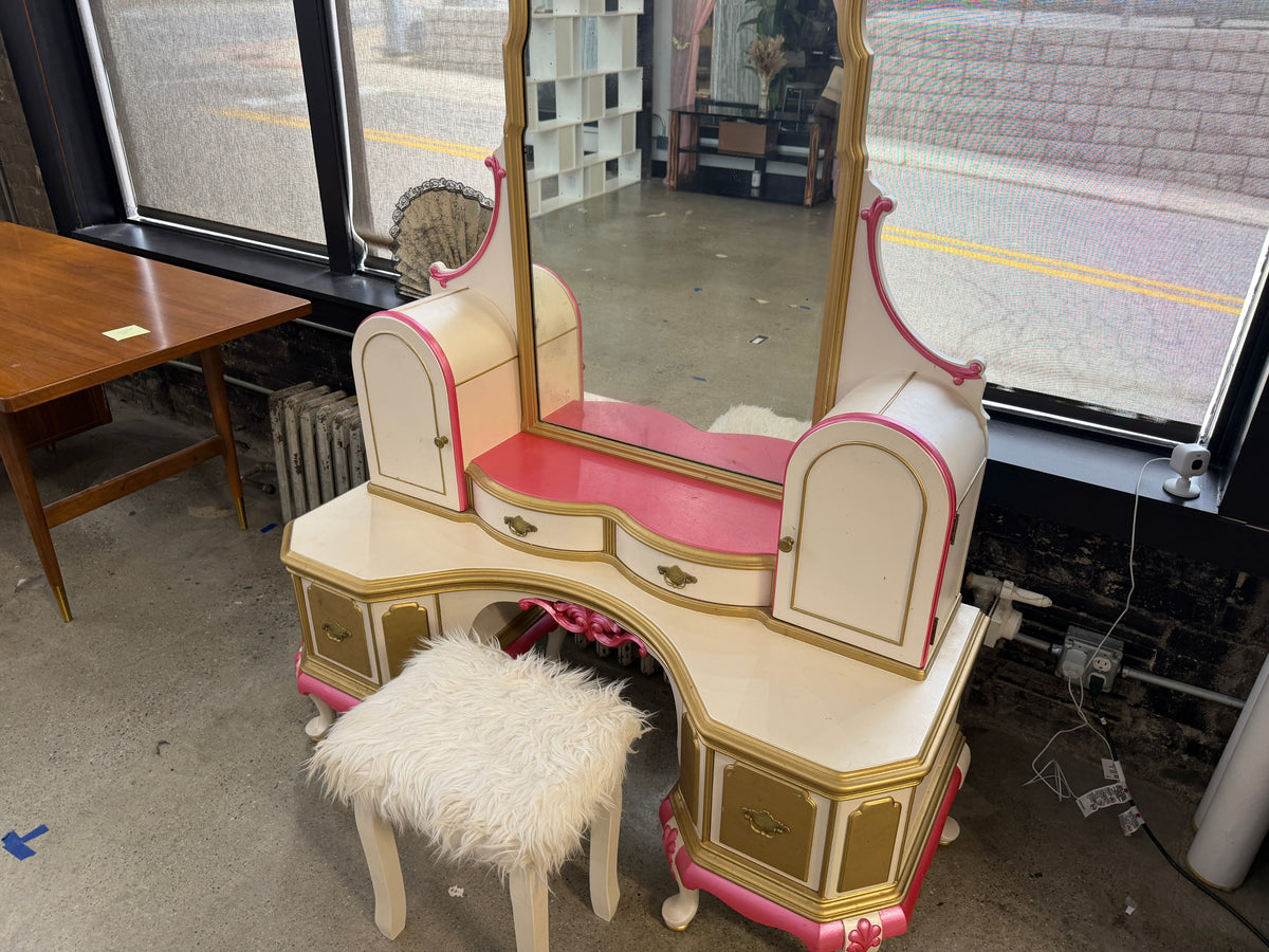 Early 1900s restored French Provincial vanity with tall center mirror, curved serpentine body, pink velvet dressing surface, gold trim, arched side storage cabinets, multiple drawers, and matching fluffy white stool — premium restored antique statement piece on consignment at The Foundry by History Loves Company in downtown Pontiac, Michigan