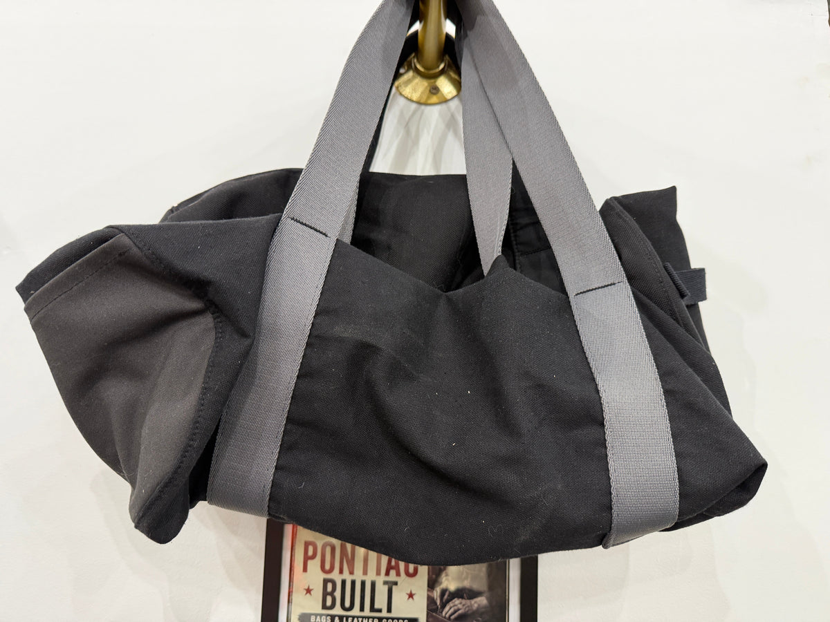 The Overnight Duffle in black with grey straps by The Foundry, Pontiac Built sign in background