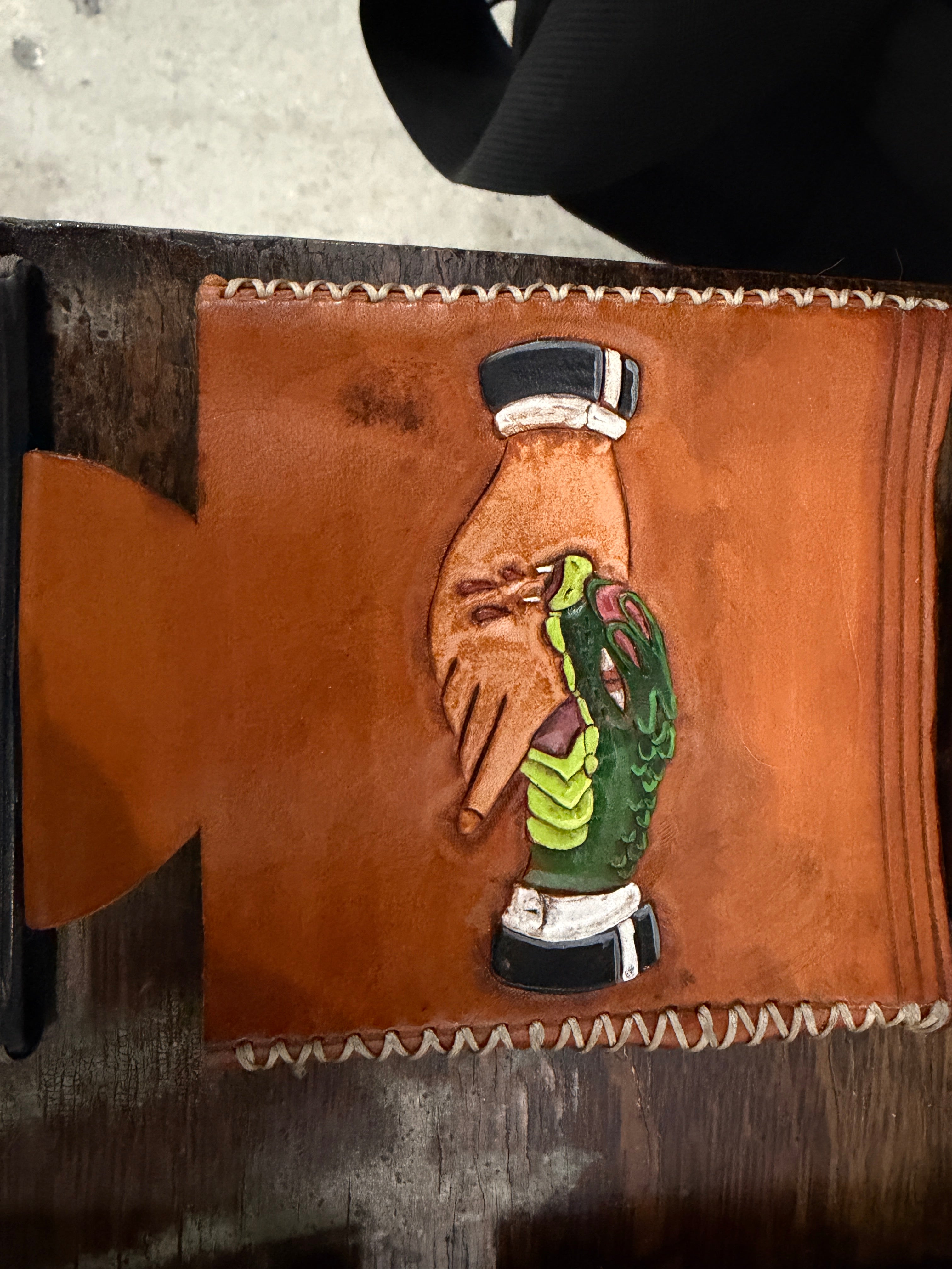 The Foundry Can Sleeve in cognac leather, hand-tooled and painted joint design — hand holding lit joint with green sleeve and black cuffs, cream saddle stitch