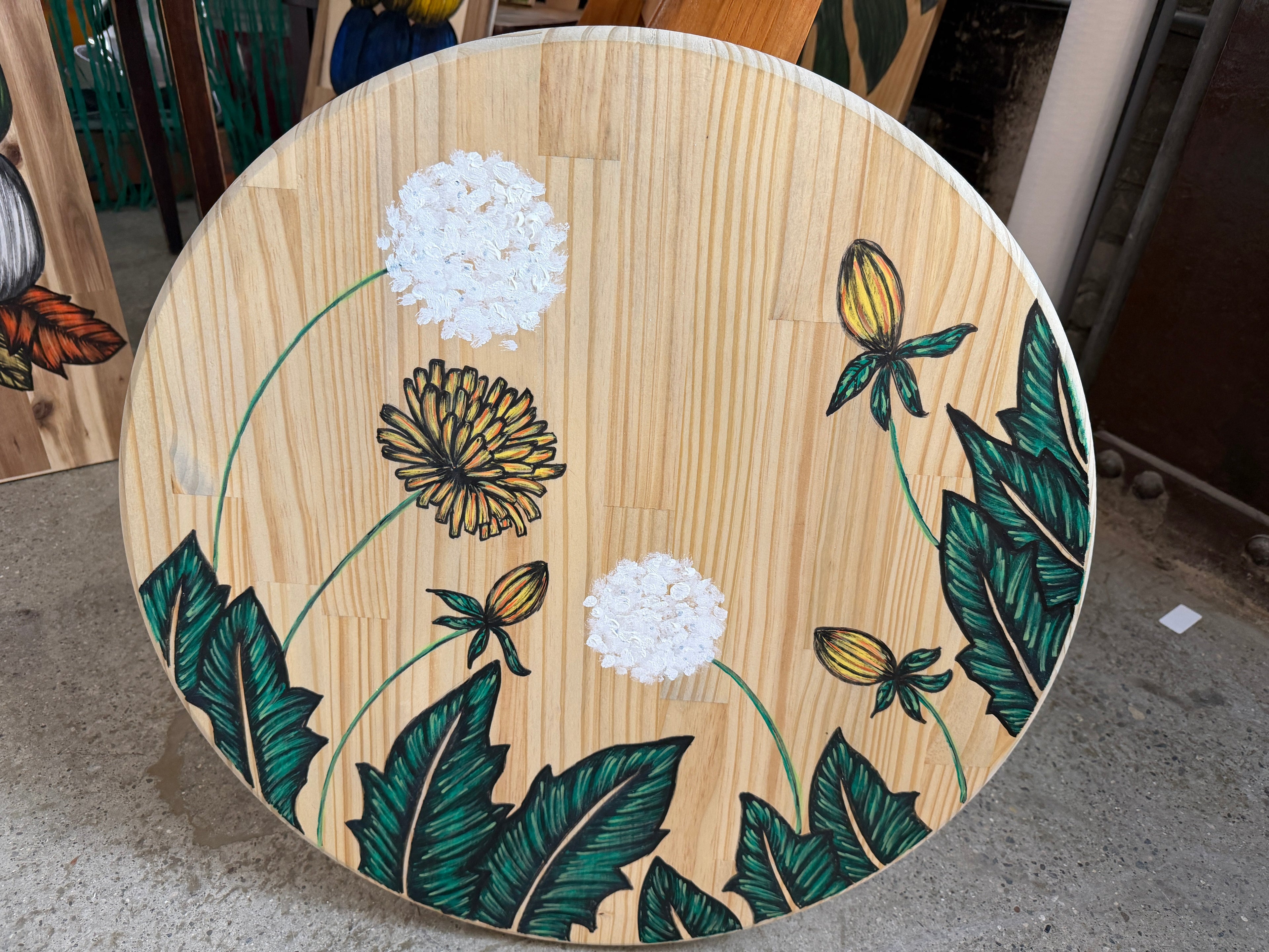 24-inch round wood panel with hand-drawn dandelions, white puffballs, yellow flowers and green tropical leaves by Renee Reese